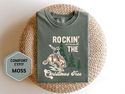 a green t-shirt with a graphic design depicting a cowboy on a horse, along with the text "ROCKIN' around THE CHRISTMAS Tree" and "COMFORT C1717 MOSS".