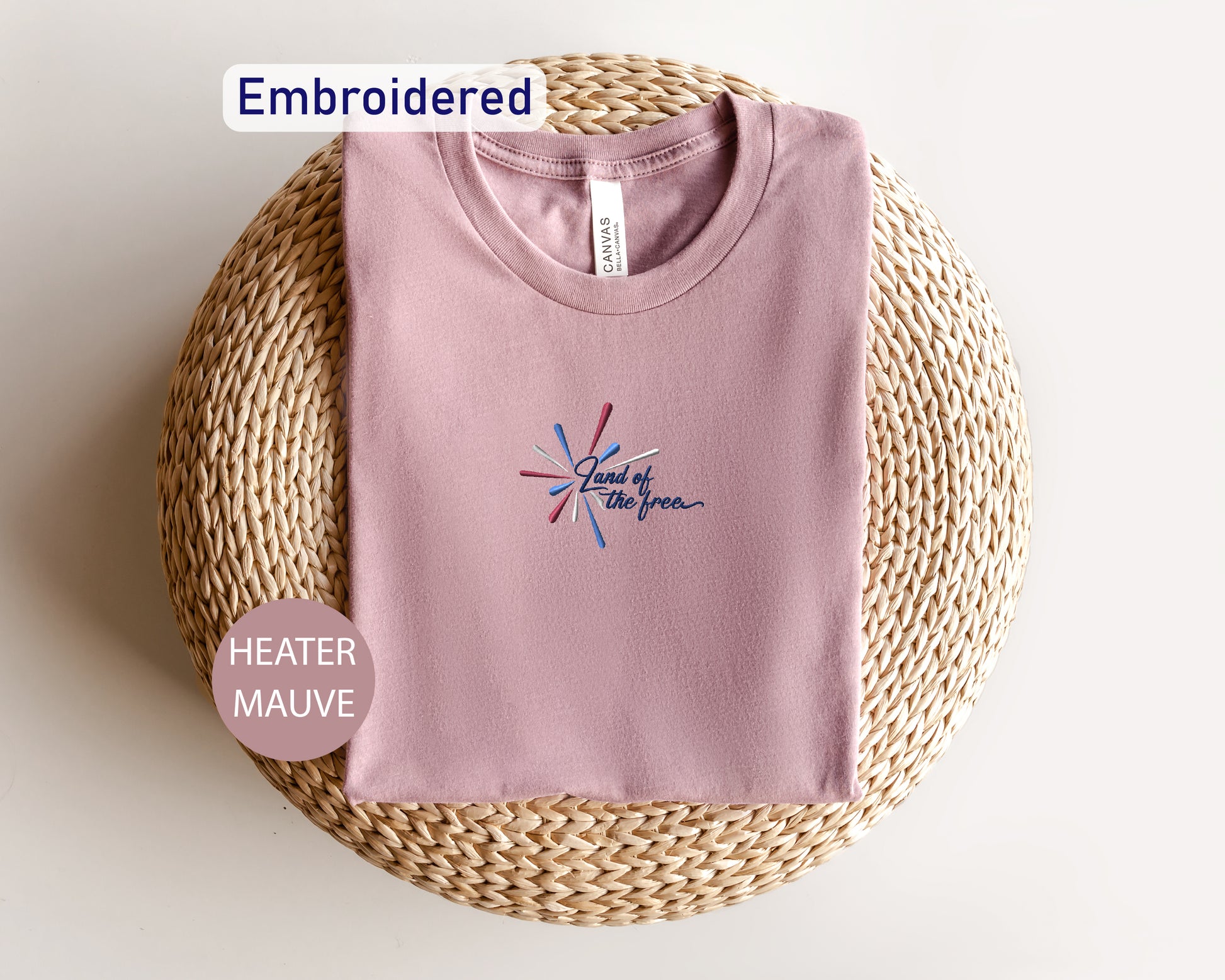 a pink t-shirt with a graphic design, laid out on a woven mat or basket. The t-shirt is embroidered and appears to be a part of a collection.