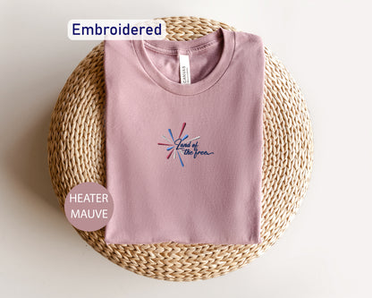 a pink t-shirt with a graphic design, laid out on a woven mat or basket. The t-shirt is embroidered and appears to be a part of a collection.