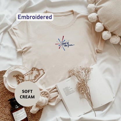a beige t-shirt with a colorful embroidered design, a bottle of soft cream, a book, and some dried flowers on a white background.