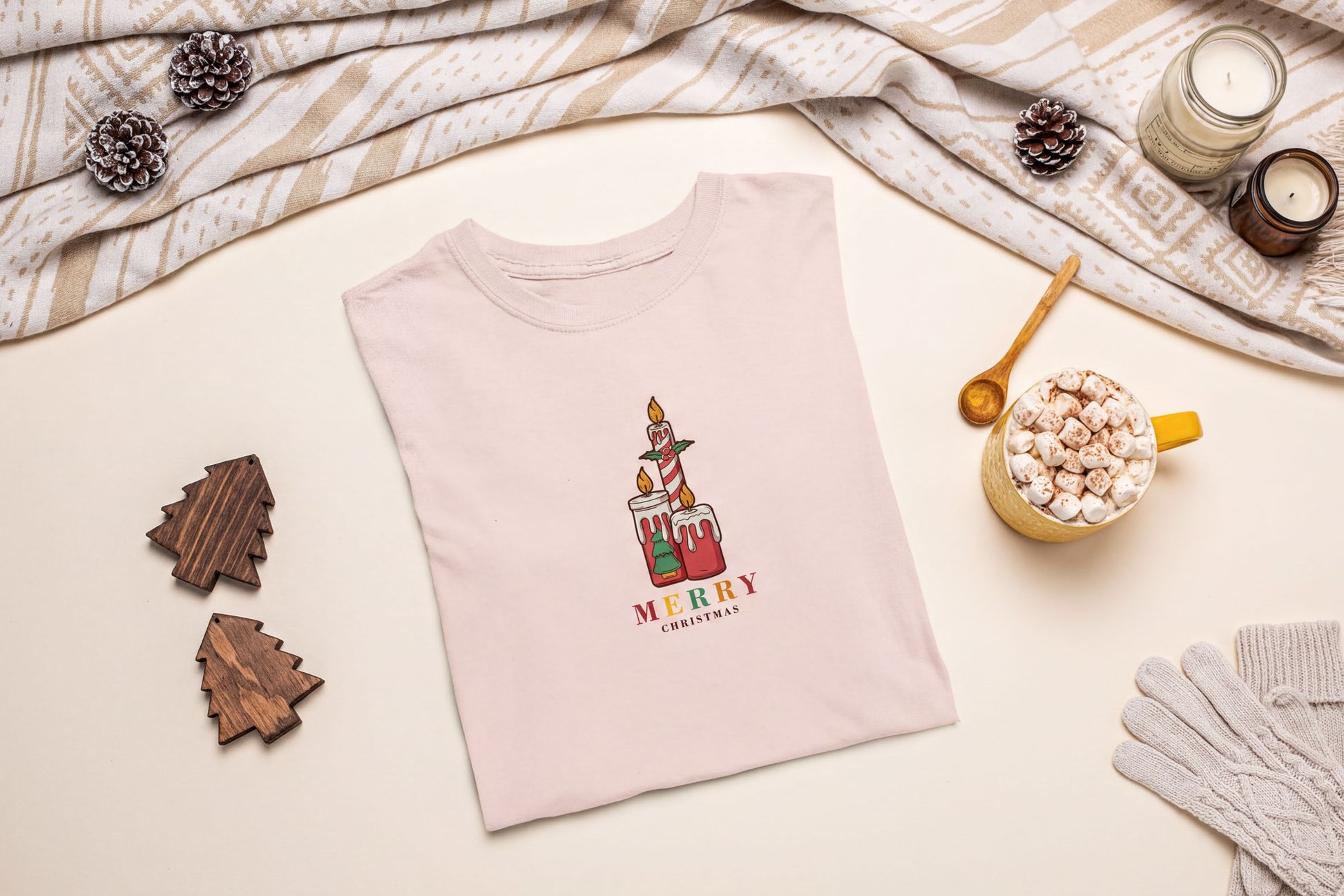 A pink t-shirt with a Christmas-themed graphic, a wooden candle holder, a mug of hot chocolate, and some pine cones are arranged on a beige background.