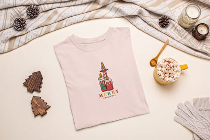 A pink t-shirt with a Christmas-themed graphic, a wooden candle holder, a mug of hot chocolate, and some pine cones are arranged on a beige background.