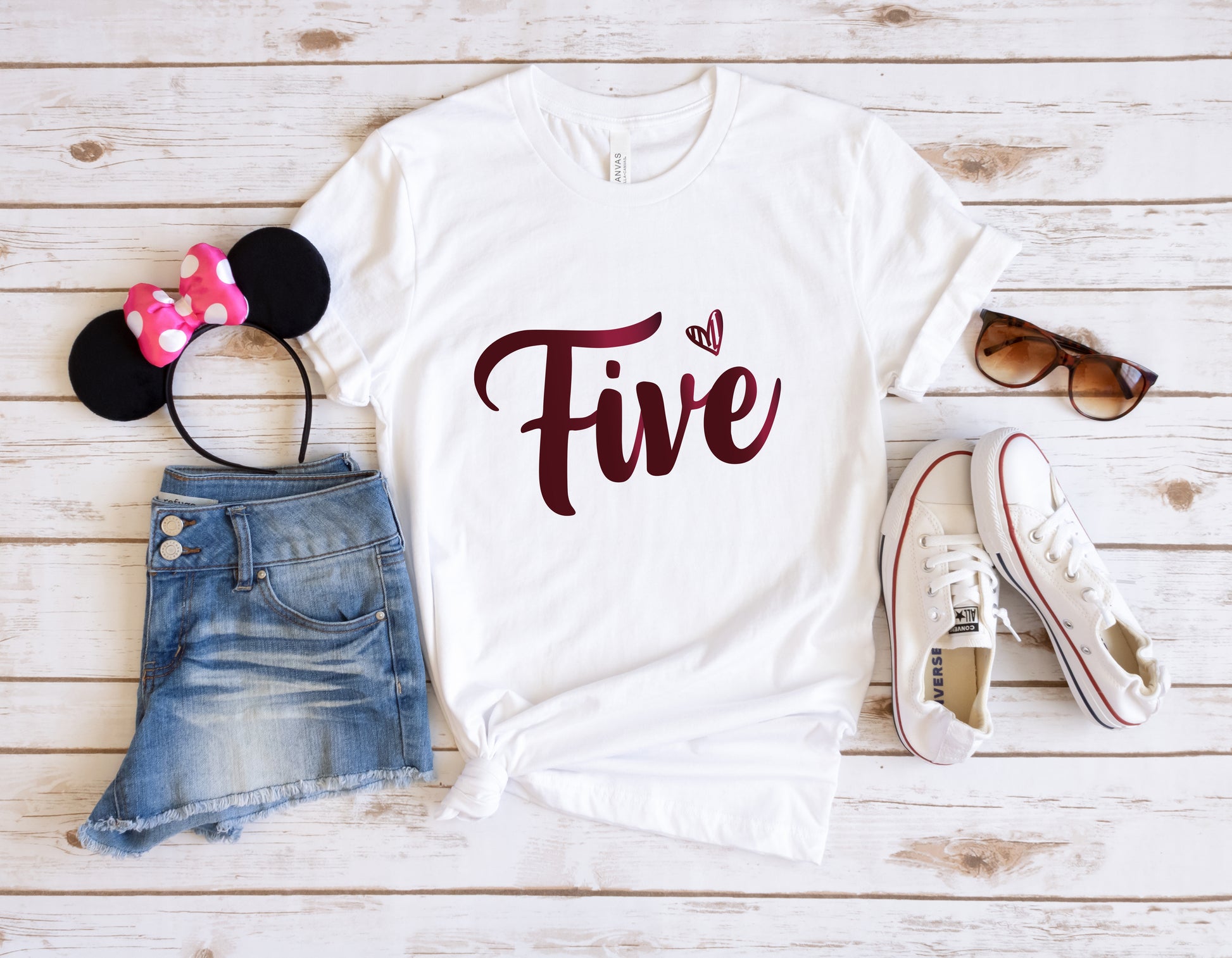 A casual outfit consisting of a white t-shirt with the number "Five" printed on it, a pair of denim shorts, a pair of sunglasses, and a pair of white sneakers is laid out on a wooden surface.