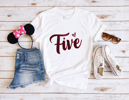 A casual outfit consisting of a white t-shirt with the number "Five" printed on it, a pair of denim shorts, a pair of sunglasses, and a pair of white sneakers is laid out on a wooden surface.