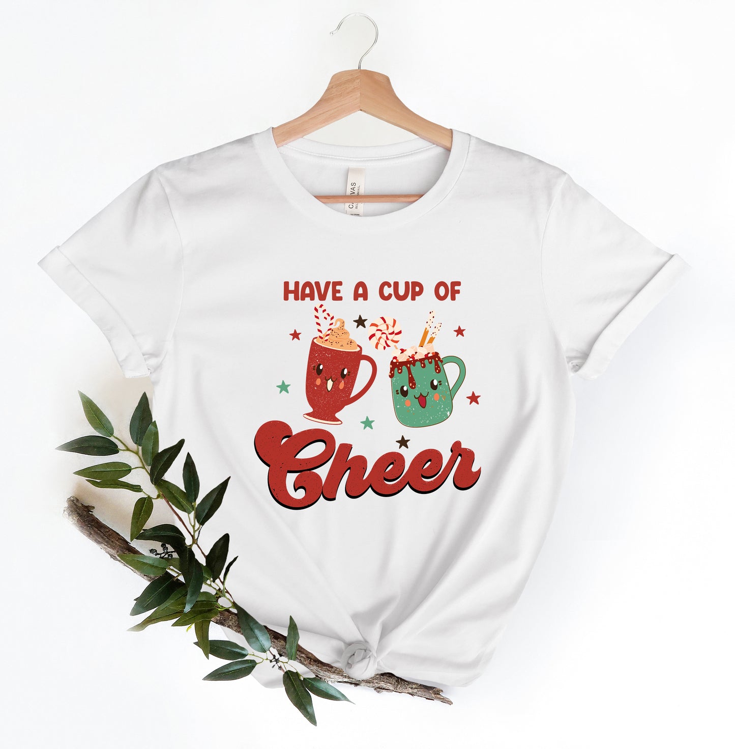 A white t-shirt with a graphic design featuring two colorful mugs with smiling faces, surrounded by stars and the text "Have a cup of cheer".