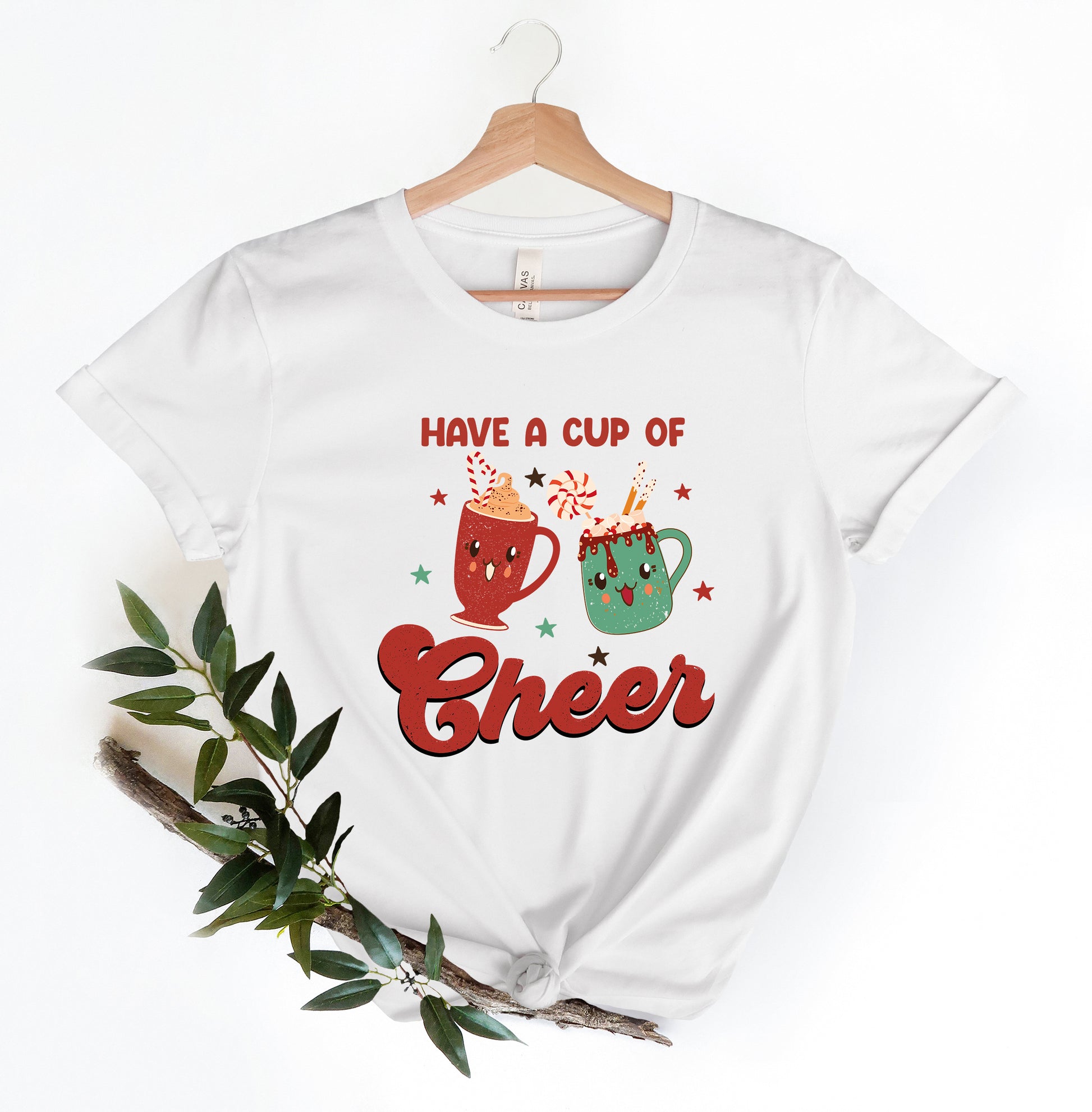 A white t-shirt with a graphic design featuring two colorful mugs with smiling faces, surrounded by stars and the text "Have a cup of cheer".