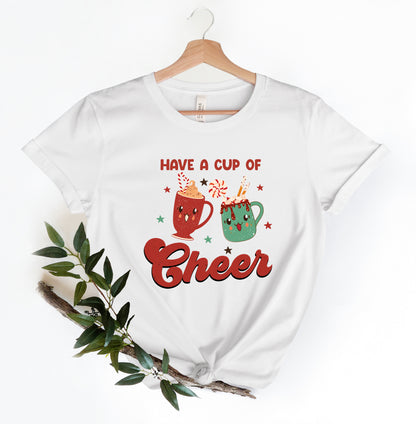 A white t-shirt with a graphic design featuring two colorful mugs with smiling faces, surrounded by stars and the text "Have a cup of cheer".