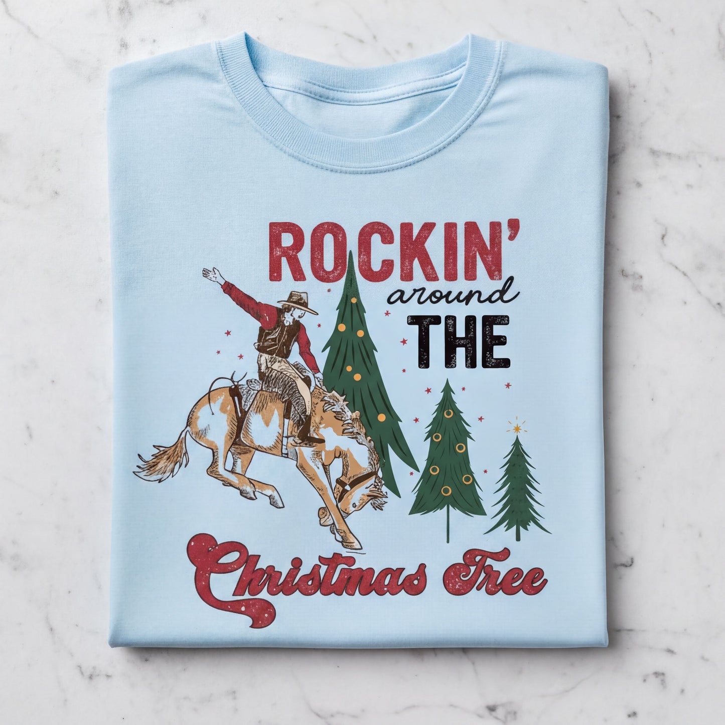 A light blue t-shirt with a graphic design depicting a cowboy riding a horse, with Christmas trees and the text "ROCKIN' around THE CHRISTMAS FREE" printed on it.