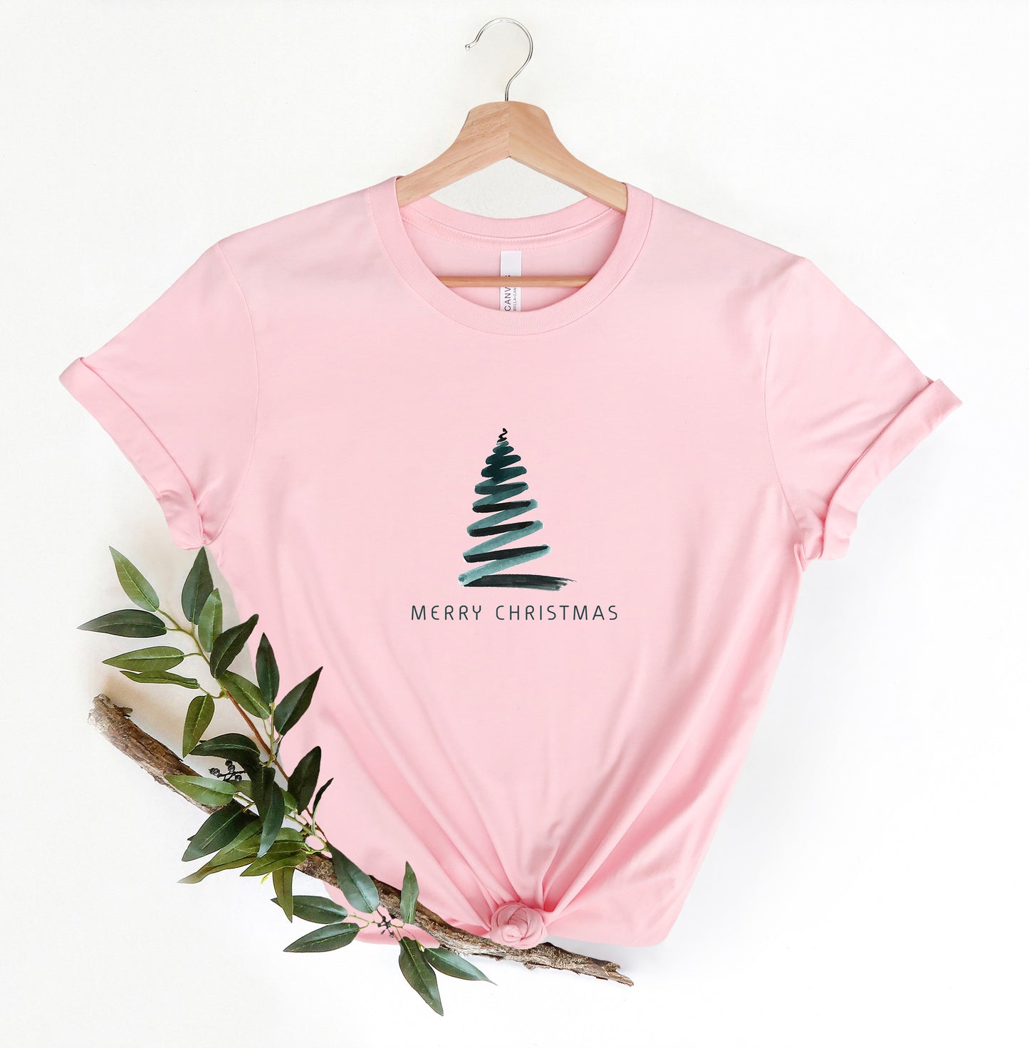 A pink t-shirt with a Christmas tree graphic and the text "Merry Christmas" printed on it, hanging on a wooden hanger.