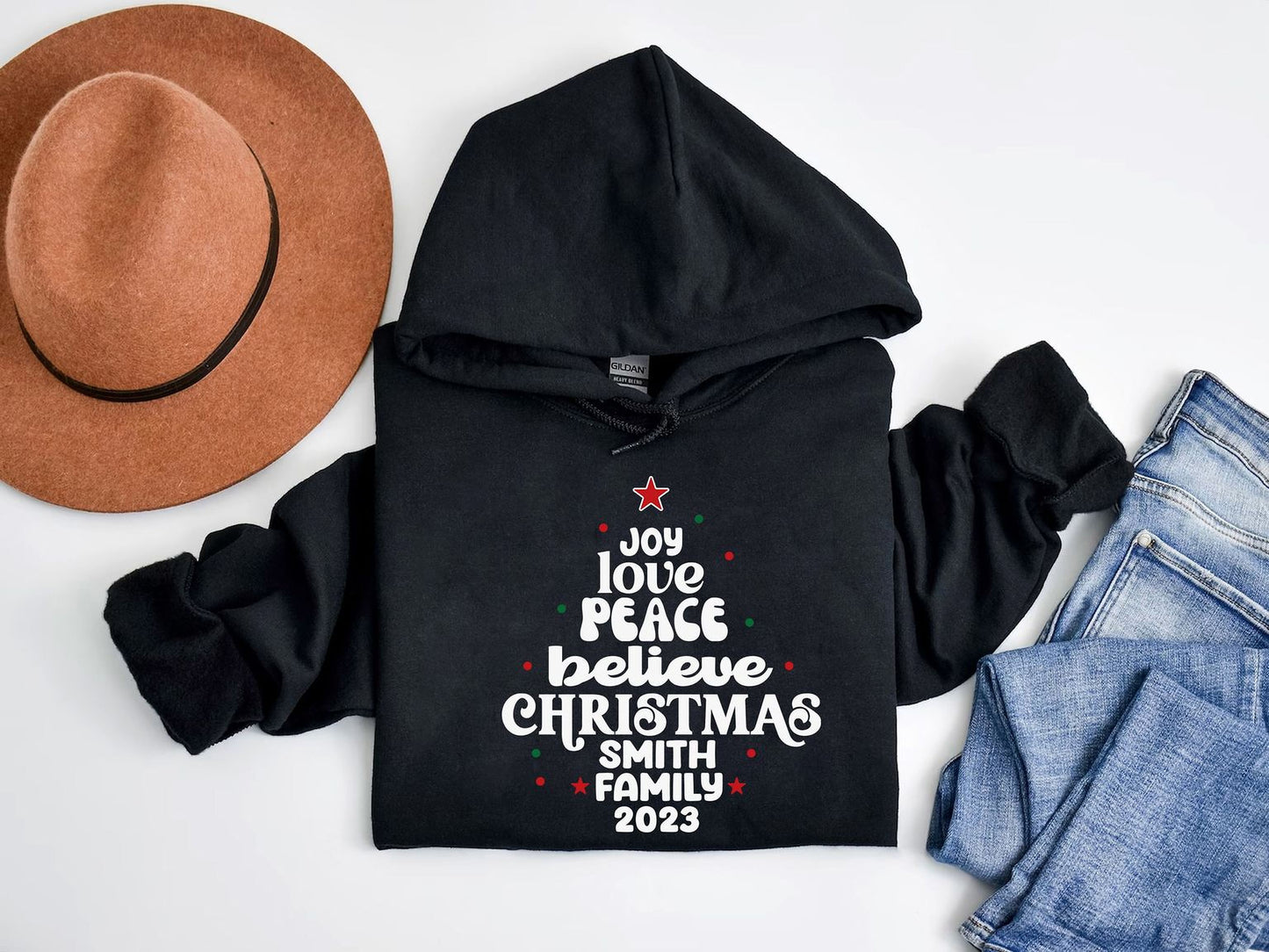 A black hoodie with a Christmas tree design and text, a brown hat, and a pair of blue jeans are arranged on a white background.