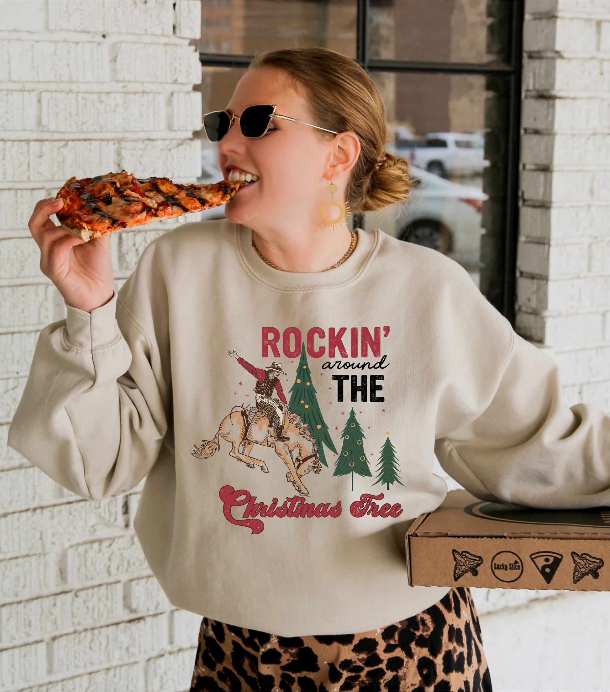 A woman wearing a sweater with a graphic design of a cowboy on a horse and the text "ROCKIN' around THE CHRISTMAS Tree" is eating a slice of pizza.