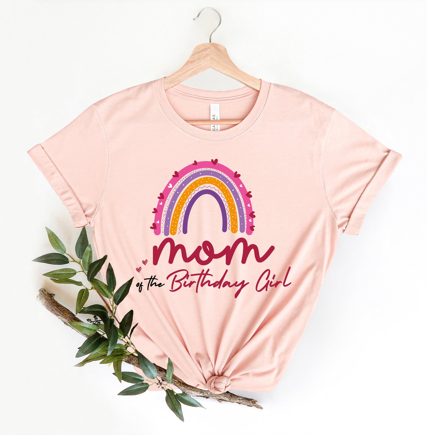 A pink t-shirt with a rainbow graphic and the text "Mom of the Birthday Girl" printed on it, hanging on a wooden hanger.