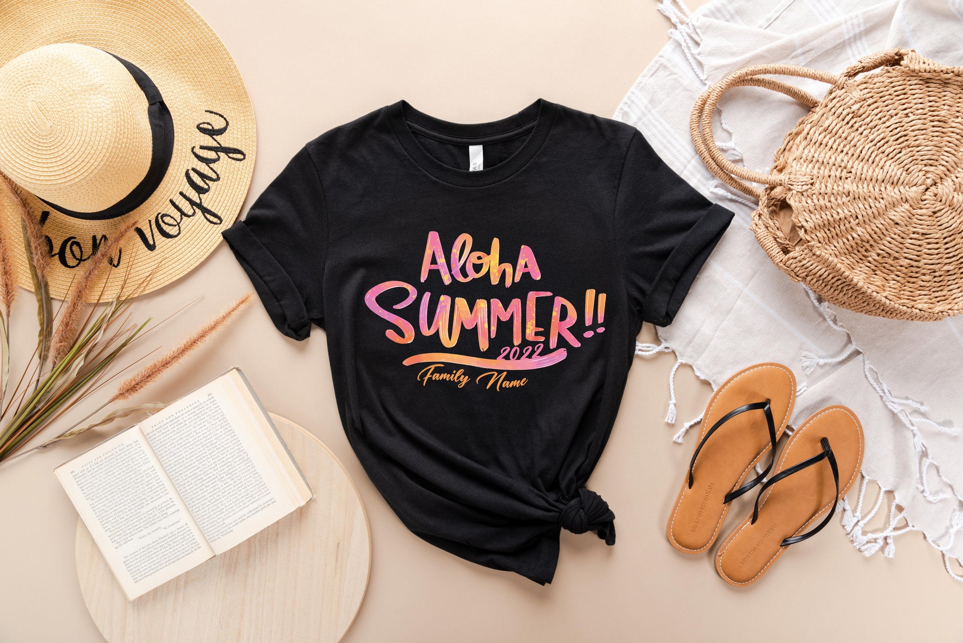 A casual summer outfit consisting of a black t-shirt with a colorful text design, a straw hat, a woven basket, a book, and sandals is arranged on a beige background.