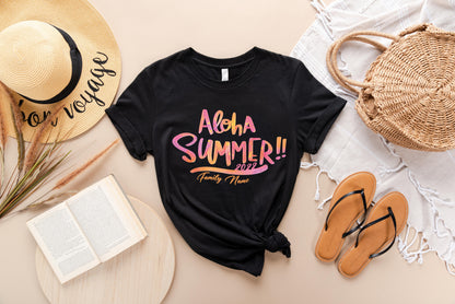 A casual summer outfit consisting of a black t-shirt with a colorful text design, a straw hat, a woven basket, a book, and sandals is arranged on a beige background.