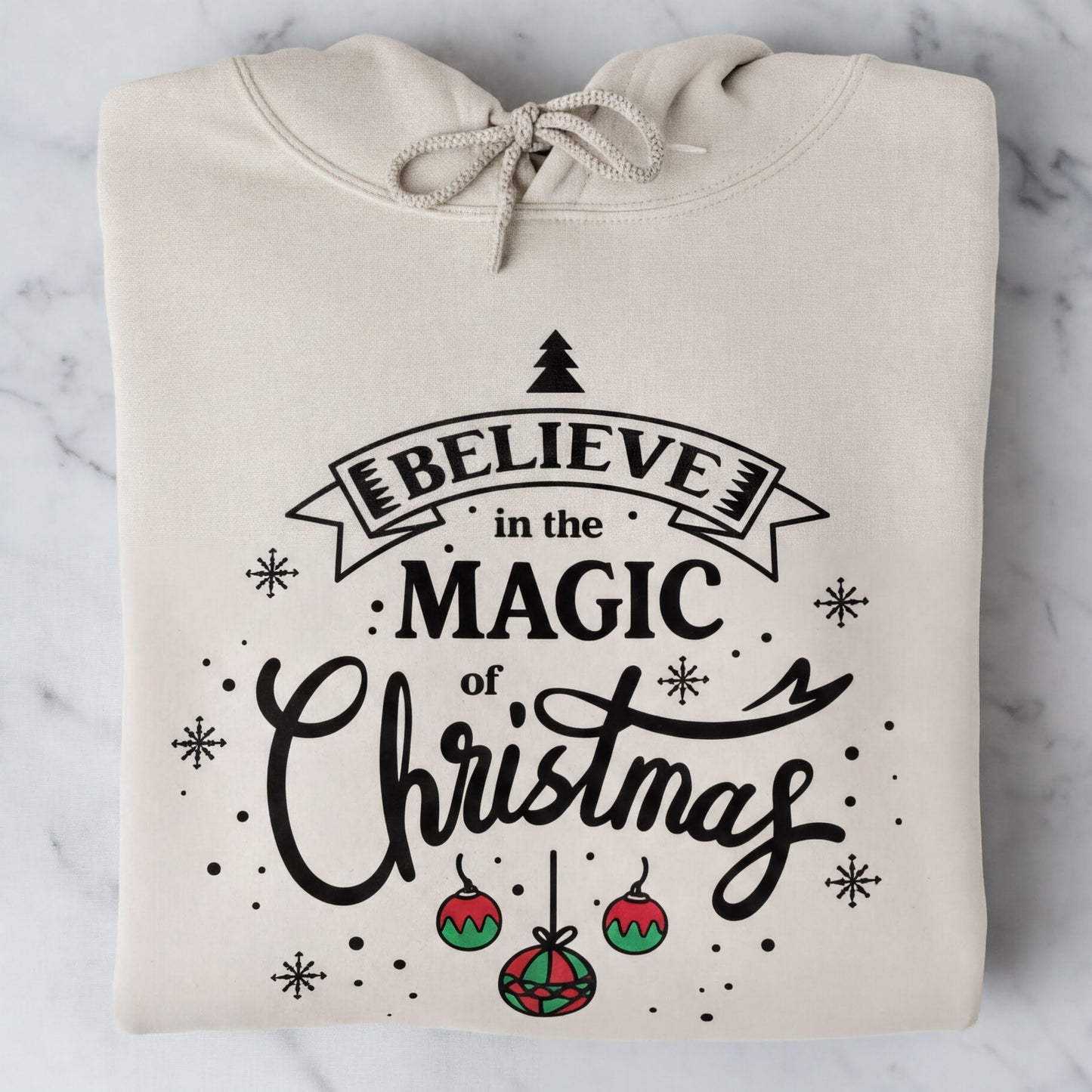 A beige hoodie with a printed design that says "Believe in the Magic of Christmas" and includes Christmas-themed elements such as snowflakes and ornaments.