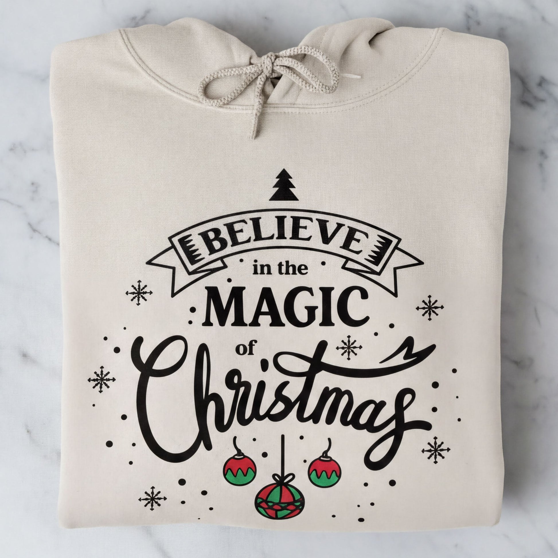 A beige hoodie with a printed design that says "Believe in the Magic of Christmas" and includes Christmas-themed elements such as snowflakes and ornaments.