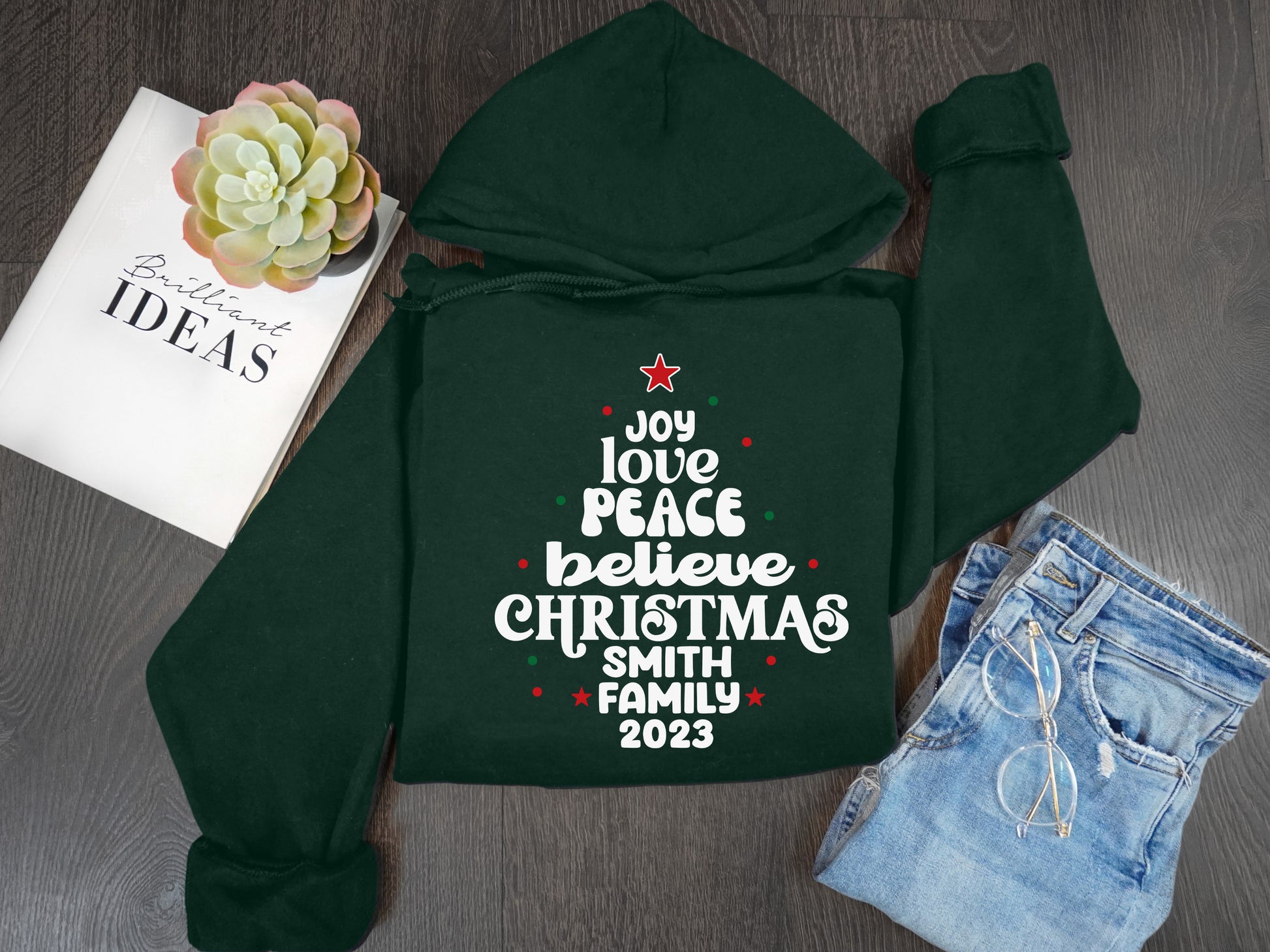 A green hoodie with a Christmas tree design and text, a pair of jeans, a book titled "Brilliant Ideas", and a succulent plant are arranged on a wooden surface.