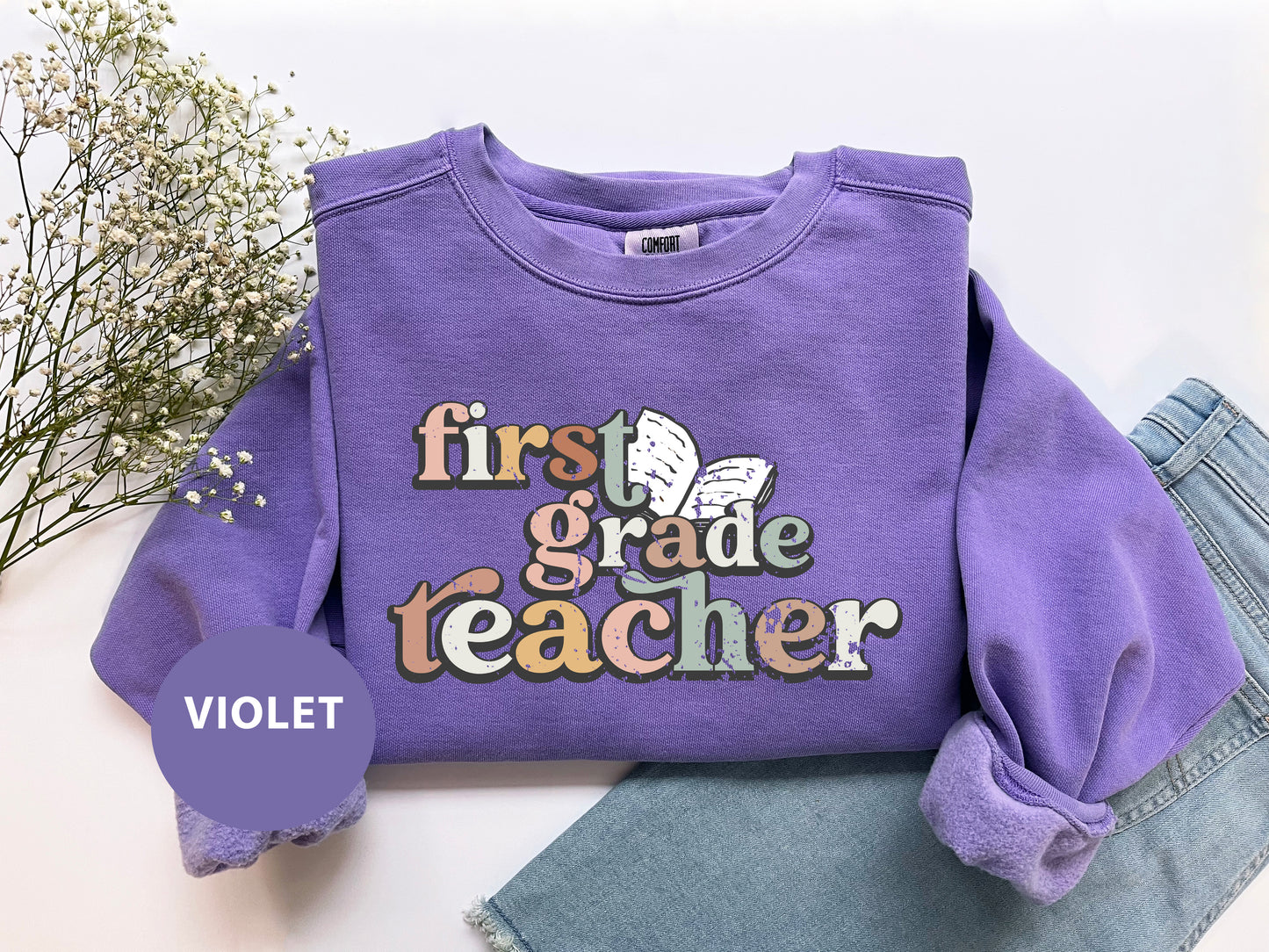 a purple sweatshirt with the text "first grade teacher" printed on it, along with a floral design. The sweatshirt is laid out on a white surface, with a pair of jeans visible underneath it.
