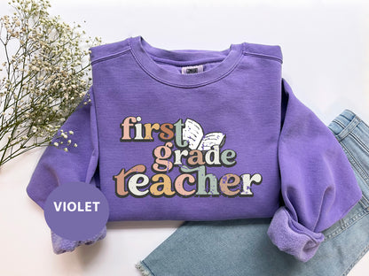 a purple sweatshirt with the text "first grade teacher" printed on it, along with a floral design. The sweatshirt is laid out on a white surface, with a pair of jeans visible underneath it.